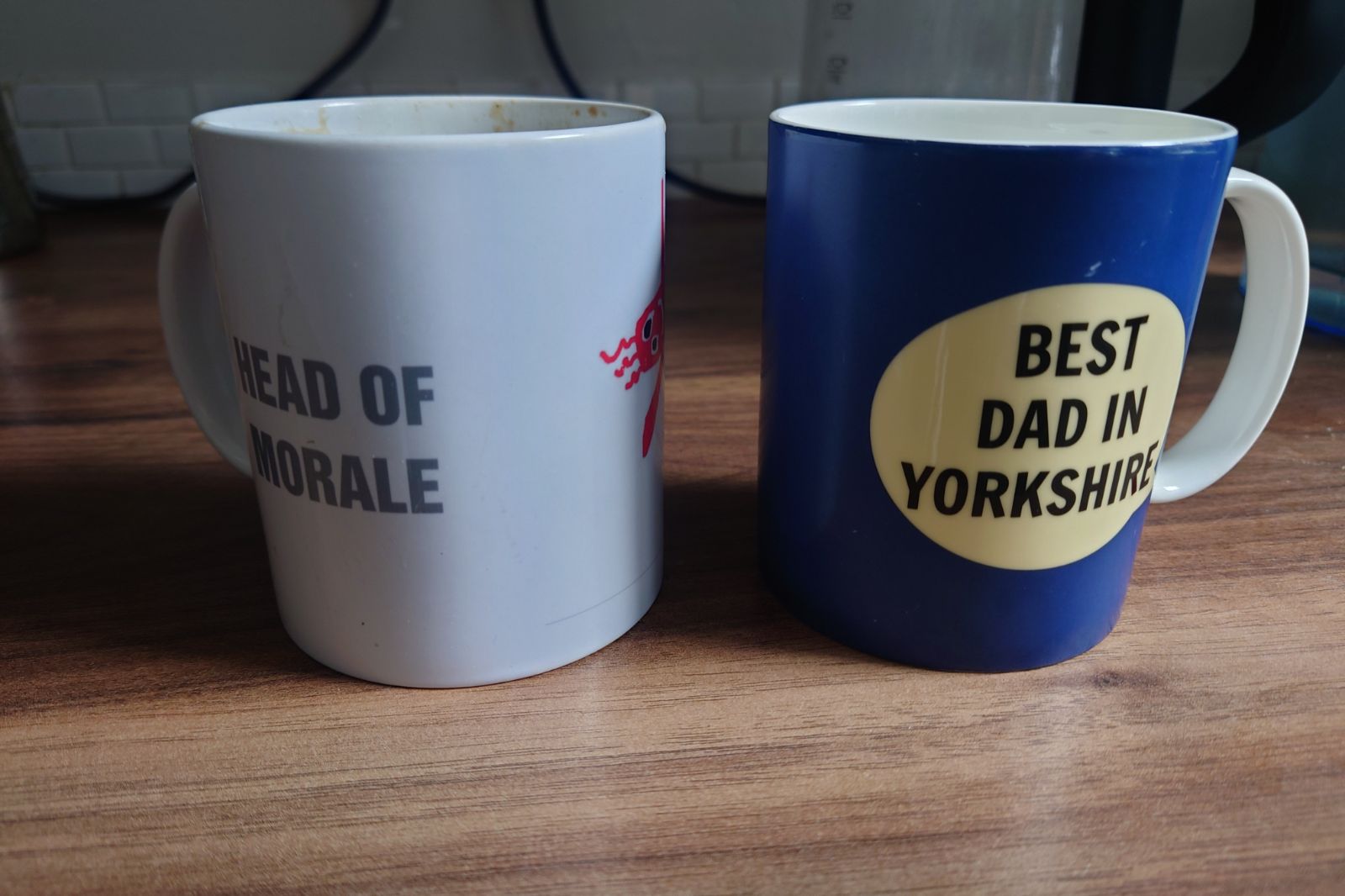 Two mugs on a dark brown wood kitchen top. On the left is a white mug with grey capitalised text that reads 'HEAD OF MORALE'. On the right is a blue mug, with black text in a pale yellow circle that reads 'BEST DAD IN YORKSHIRE'.