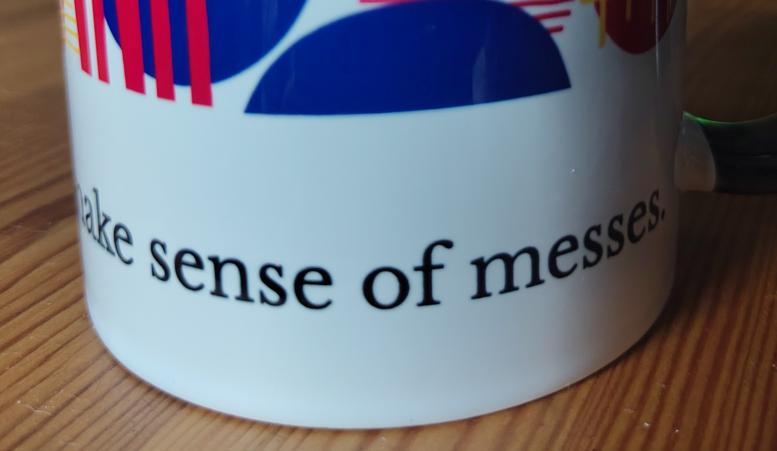 Closeup of the bottom half of a white mug on a wooden kitchen table. On the mug is the black text 'I make sense of messes'.