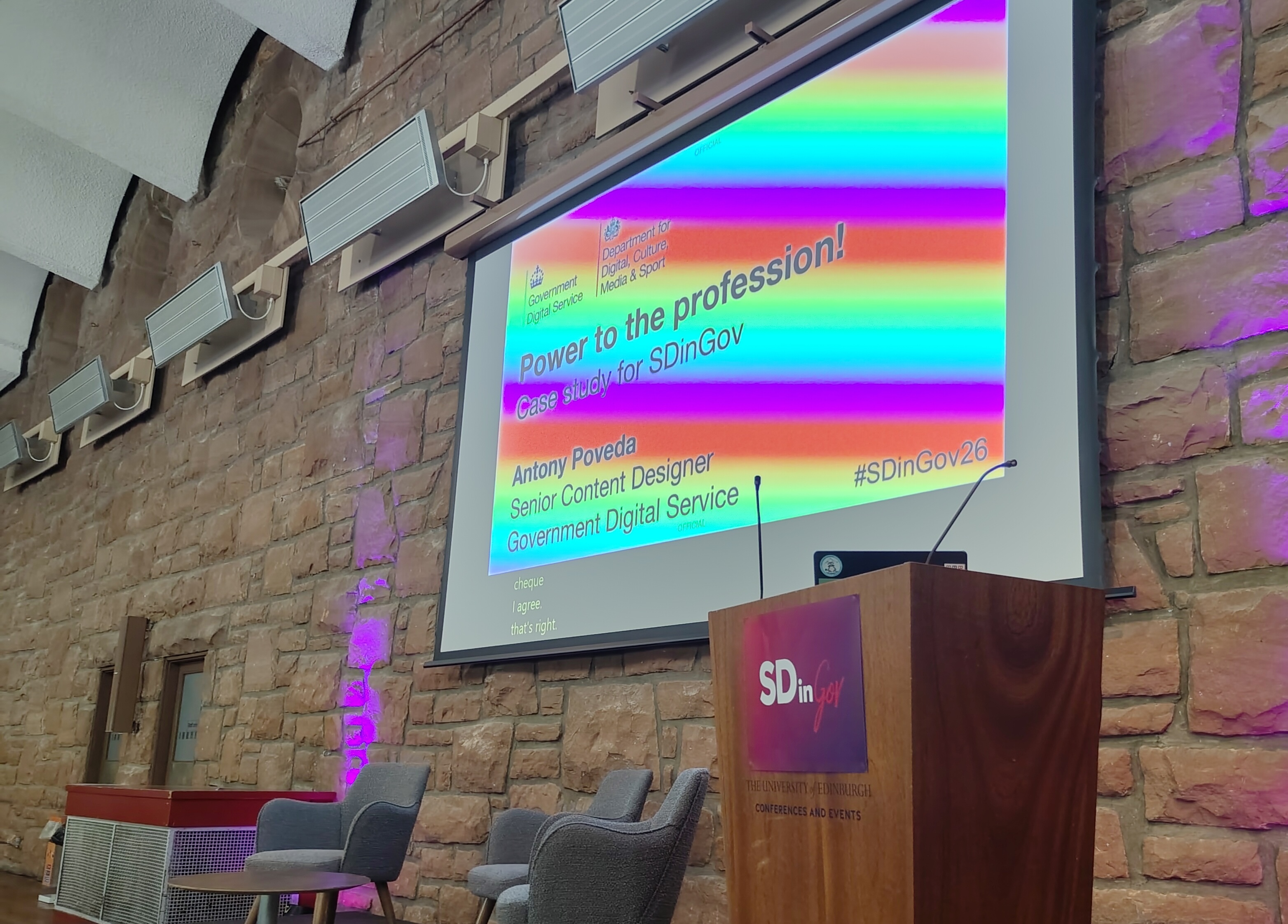 Photo of an empty conference stage with three chairs and a wooden lectern, taken from stage right. A projected slide says 'Power to the Profession! Case Study, Antony Poveda, Senior Content Designer, Government Digital Service'. The slide appears to have a rainbow pattern due to the camera. The lectern is on the right and displays the logo for 'SDinGov'.